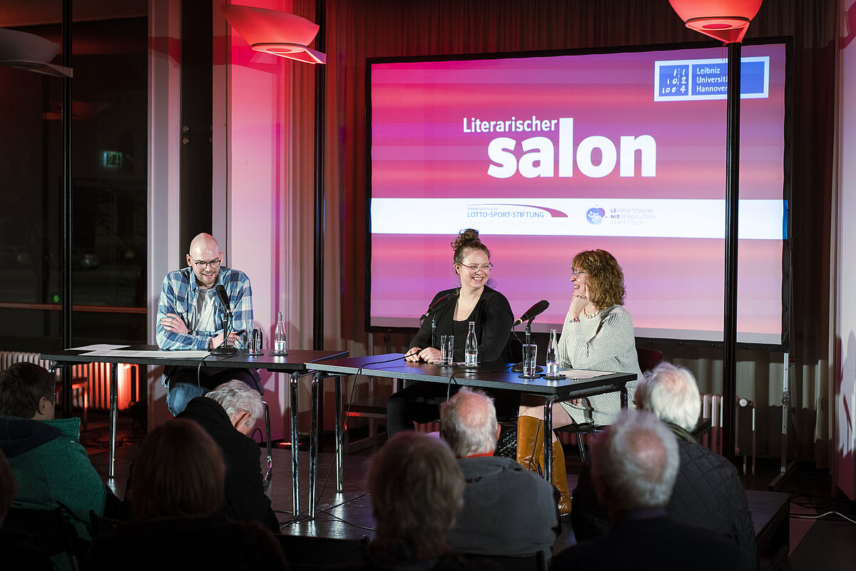Vor dem pinken Hintergrund des literarischen Salons sitzt der Moderator mit den zwei Gästinnen.