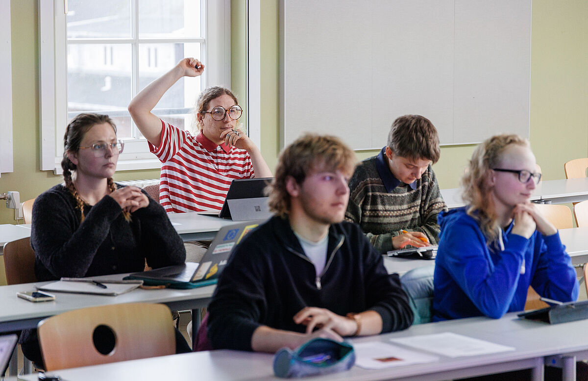 Fünf Studierende sitzen im Seminar und schauen interessiert nach vorne. Hinten meldet sich eine Person.