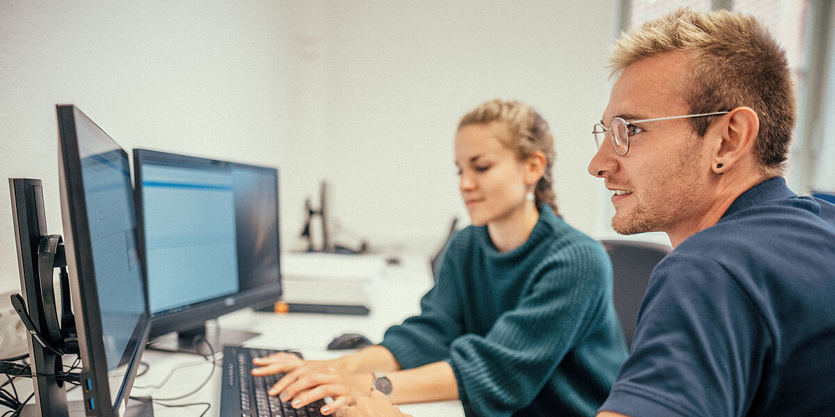 Eine Studentin und ein Student arbeiten gemeinsam im Sprachenzentrum an einem Notebook mit zwei Bildschirmen.