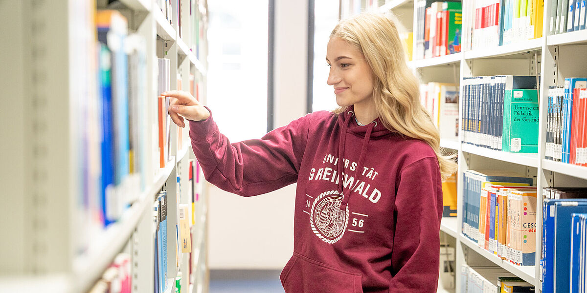 Studierende in der Bibliothek: Eine Studentin wählt in einer Bibliothek ein Buch aus einem Regal.