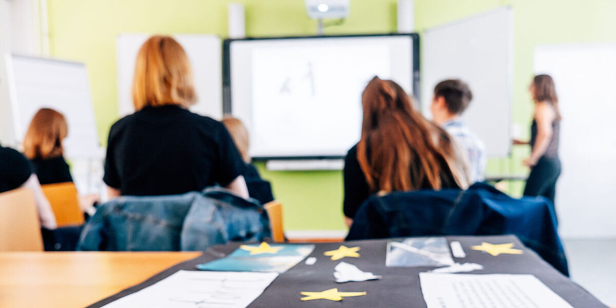 Eine Nahaufnahme eines dunklen Plakats im Unterricht ist zu sehen. Im Hintergrund sieht man Schüler von hinten in einem Klassenraum sitzen.