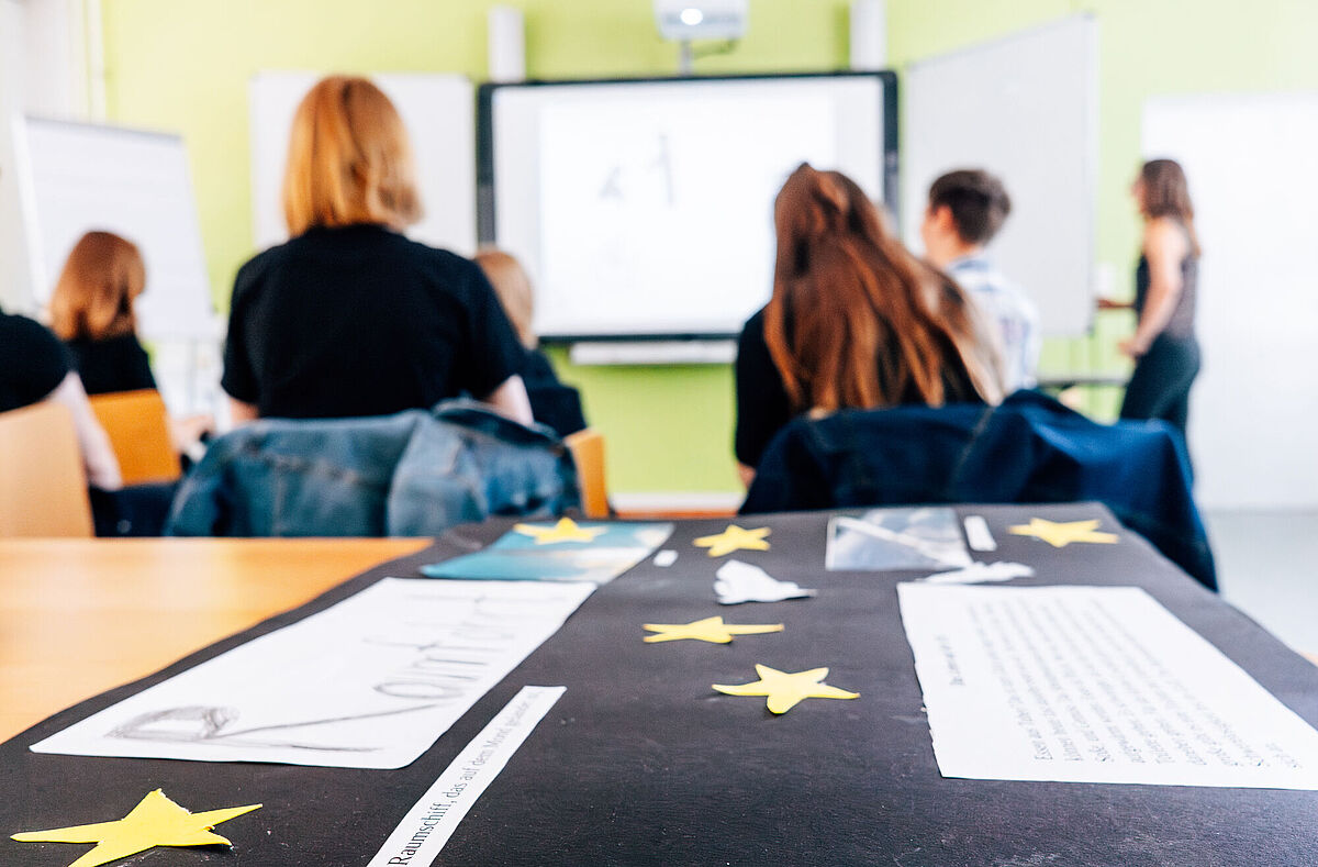 Eine Nahaufnahme eines dunklen Plakats im Unterricht ist zu sehen. Im Hintergrund sieht man Schüler von hinten in einem Klassenraum sitzen.