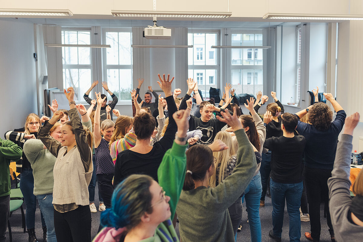 Die Gruppe steht im Raum und hebt die Hände nach oben. Es wird eine unterschiedliche Anzahl an Fingern gezeigt.