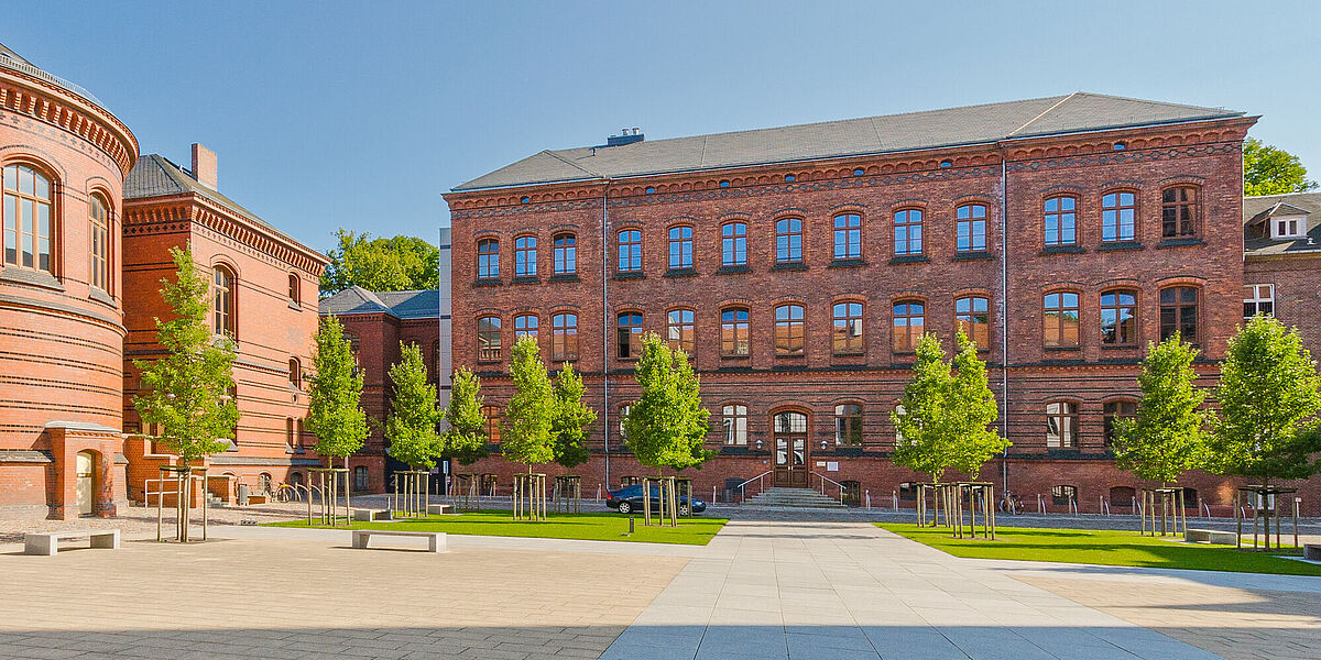 Außenansicht der backsteinernen ehemaligen Augenklinik hinter dem begrünten und sonnenbeschienen Innenhof des Historischen Campus vor blauem Himmel.