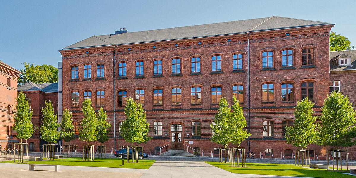 Außenansicht der backsteinernen ehemaligen Augenklinik hinter dem begrünten und sonnenbeschienen Innenhof des Historischen Campus vor blauem Himmel.