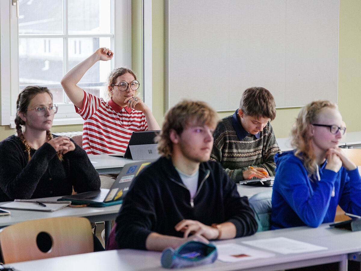 Fünf Studierende sitzen im Seminar und schauen interessiert nach vorne. Hinten meldet sich eine Person.
