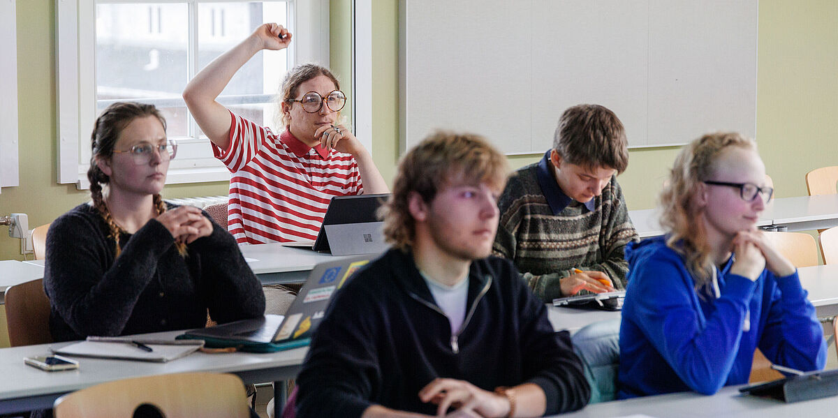 Fünf Studierende sitzen im Seminar und schauen interessiert nach vorne. Hinten meldet sich eine Person.