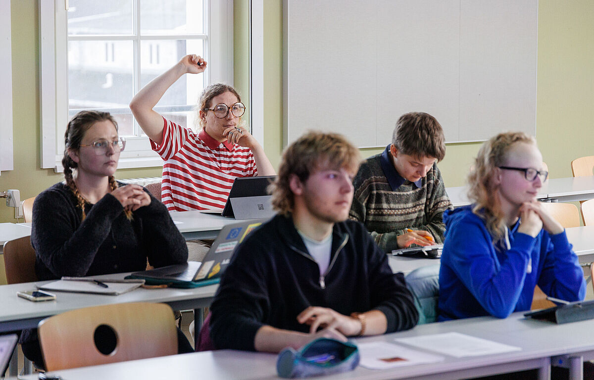Fünf Studierende sitzen im Seminar und schauen interessiert nach vorne. Hinten meldet sich eine Person.