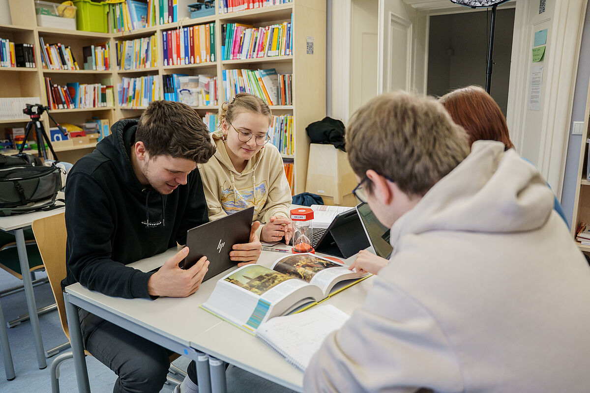 Vier Studierende sitzen an einem Tisch und lernen gemeinsam. Auf dem Tisch liegen viele Lernutensilien, wie Tablets, Bücher, Stifte, Notizhefte.
