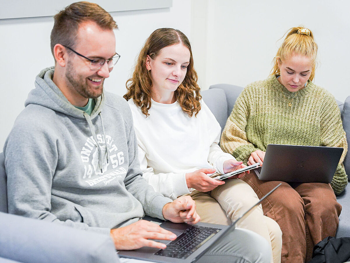 Zwei Studentinnen und ein Student sitzen beisammen und arbeiten an ihren Laptops.