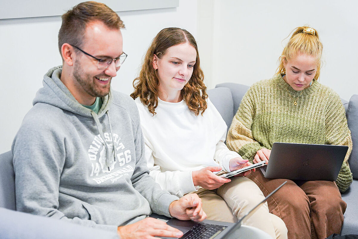 Zwei Studentinnen und ein Student sitzen beisammen und arbeiten an ihren Laptops.